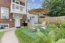 Cozy patio area with a dining set, grill, and greenery beside a brick building.