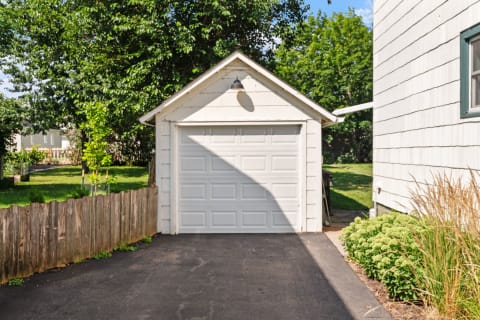 Small white garage with a closed door next to a house and lush greenery.