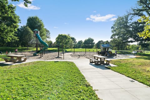 A playground with a slide, sandbox, swings, and a playhouse surrounded by green trees.