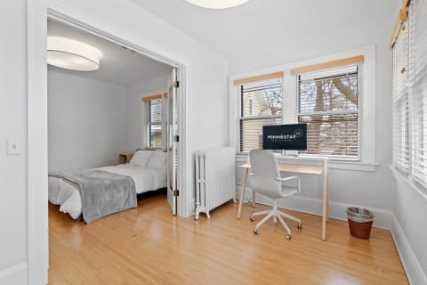 Interior view of a bedroom and workspace with a bed, desk, and light hardwood flooring.
