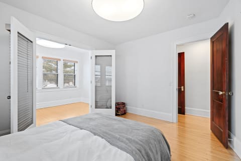 Interior view of a cozy bedroom featuring a light gray blanket on the bed, large windows, and natural wood flooring.