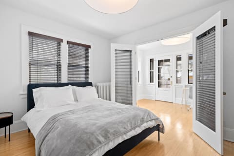 Cozy bedroom featuring a bed with white linens and a gray throw, with adjacent double doors leading to a sunlit area.