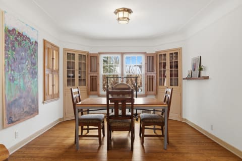 A cozy dining room with a wooden table, upholstered chairs, and large windows surrounded by glass-front cabinets.
