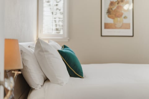 A cozy bedroom featuring white pillows and an emerald pillow on crisp linens, with framed artwork on the wall.