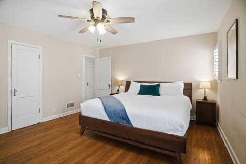 A serene bedroom with a queen bed, wooden flooring, and a ceiling fan, featuring white linens and a blue throw.