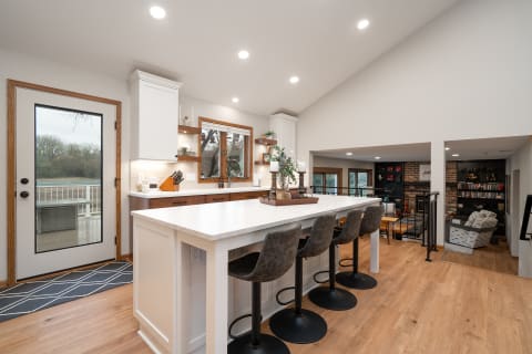 Modern kitchen with a white island, bar stools, and a view of trees outside.