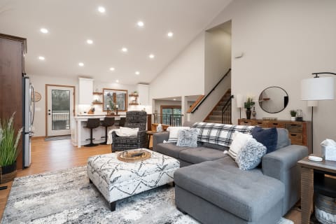 Modern living room with gray sectional sofa, patterned ottoman, and kitchen area in the background.