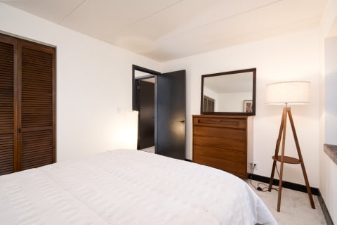 A modern bedroom with a white bed cover, wooden dresser, and floor lamp.