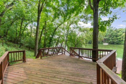 Wooden deck facing a calm lake, surrounded by trees and greenery.