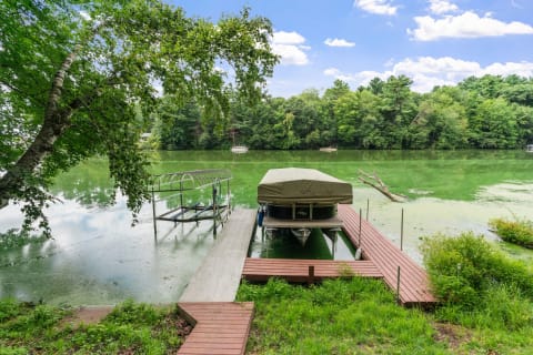 A dock with a covered boat on a green lake, surrounded by trees and under a blue sky with clouds.