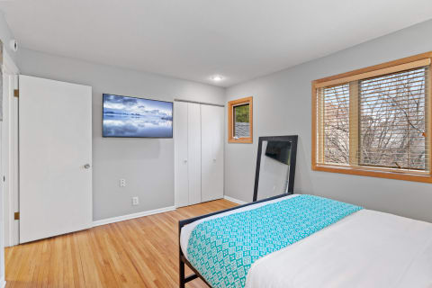 A cozy bedroom with gray walls, hardwood flooring, a turquoise bedspread, and a mounted television displaying a landscape image.