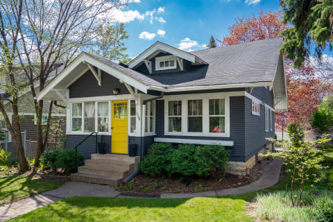 A charming gray house with a bright yellow front door surrounded by green bushes and trees.