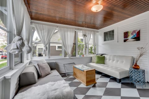 A cozy sunroom featuring grey and white sofas, a wooden coffee table, and large windows with white curtains.