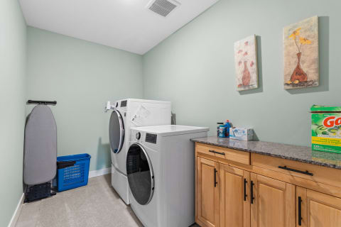 A well-organized laundry room featuring a washing machine, dryer, ironing board, and laundry supplies against a soft green background.