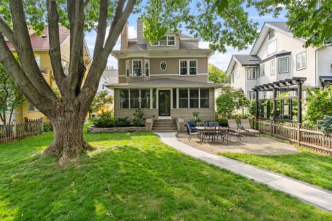 A two-story house with a large tree in the front yard and a patio area with outdoor furniture.