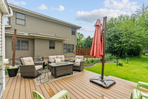 Outdoor deck featuring beige seating, a coffee table, and a coral umbrella, surrounded by greenery.
