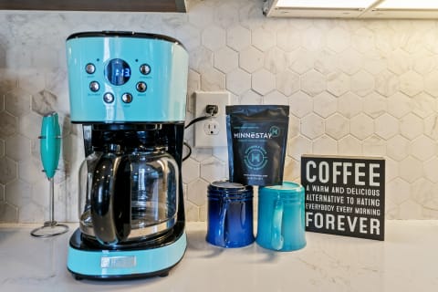 Image of a turquoise coffee maker with mugs and a humorous coffee sign on a kitchen countertop.