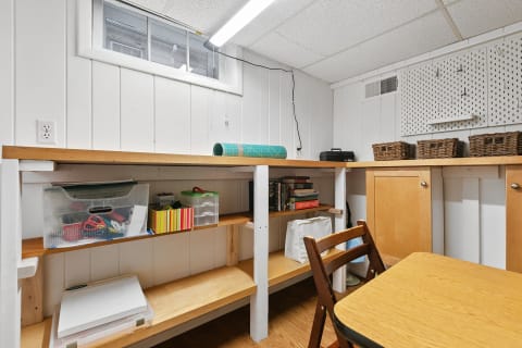A well-lit basement workspace featuring a wooden workbench, storage containers, and a small table.