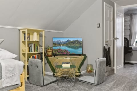 Cozy attic room with a bed, a bookshelf filled with books, a TV showing a scenic landscape, and comfortable seating.