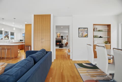 A modern open-concept living space showcasing a blue sofa, dining area with white chairs, and a kitchen with wood cabinetry.