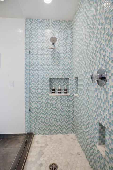 A modern shower with blue and green mosaic tiles, featuring a sleek showerhead and premium bath products on a shelf.