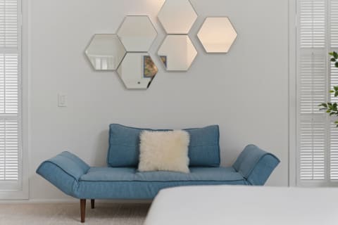 A blue daybed with a white pillow in front of hexagonal mirrors on a light wall with natural light filtering through shutters.
