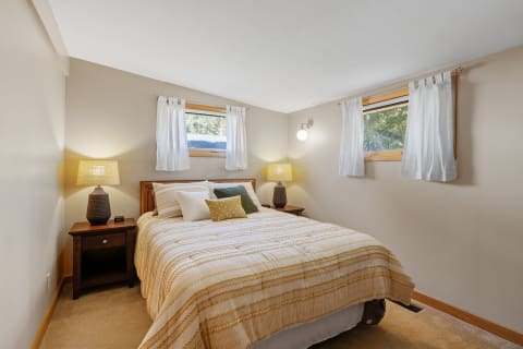 A serene bedroom featuring a bed with a striped comforter and pillows, wooden nightstands with lamps, and sheer curtains.