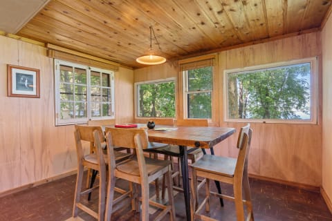 A rustic dining area with wooden walls, table, and chairs, surrounded by large windows showcasing greenery.