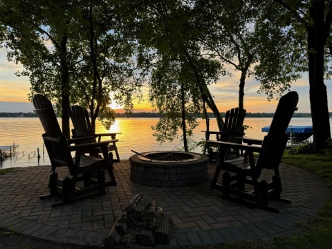 Four wooden adirondack chairs around a fire pit by a sunset lake.