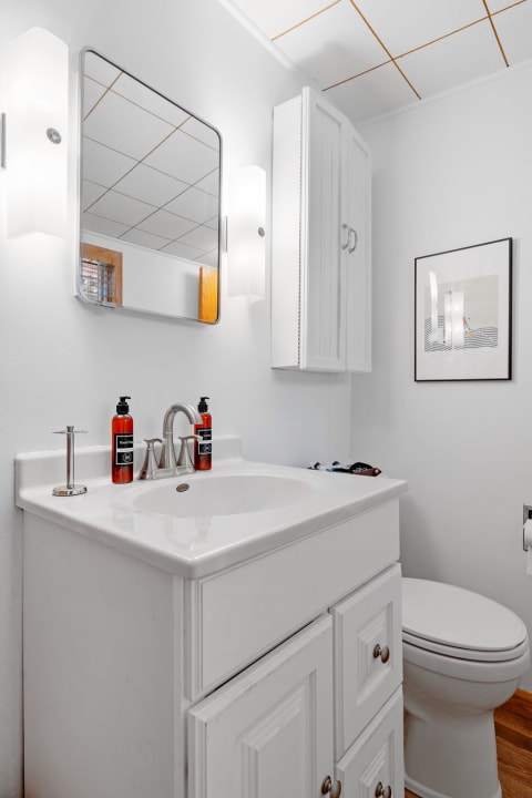 A well-lit bathroom featuring a white vanity, sink, mirror, and artwork.