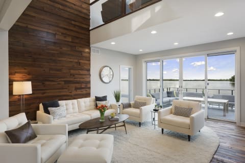 Modern living room with a wood accent wall, light furniture, and a view of a lake through glass doors.