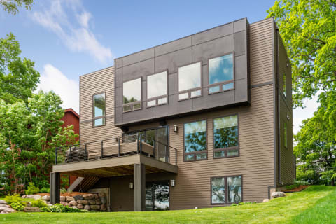 A modern house with dual levels, featuring large windows and a balcony overlooking green surroundings.