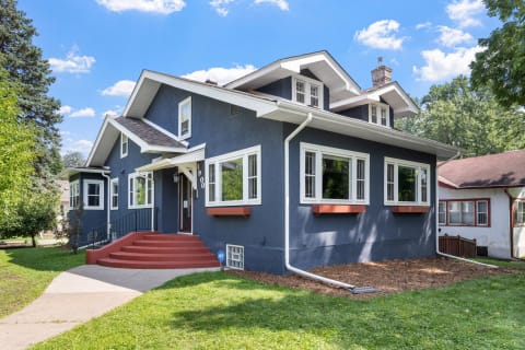 A two-story blue house with red steps and white trim, surrounded by greenery.