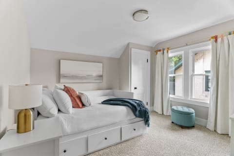 A cozy bedroom featuring a daybed, decorative pillows, and a bright window with white curtains.