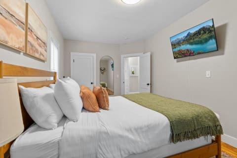 Cozy bedroom with a bed, decorative pillows, mounted TV showing a lake landscape, and beige walls.