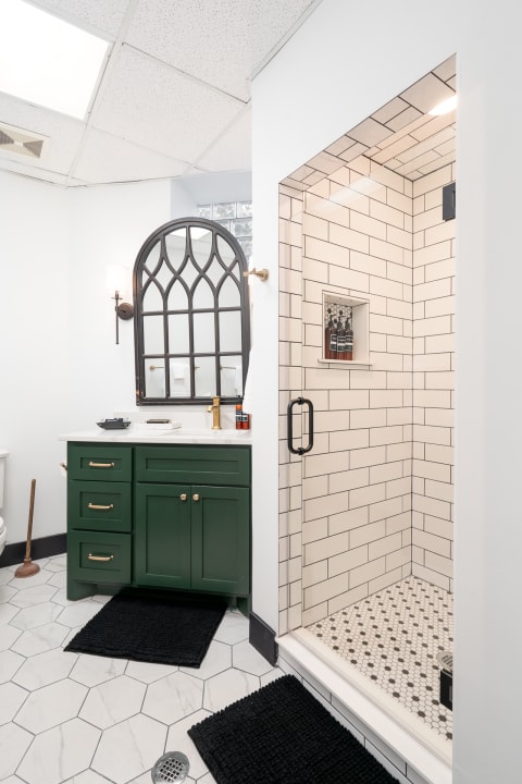 A modern bathroom with a green vanity, an arched mirror, and a subway-tiled shower.