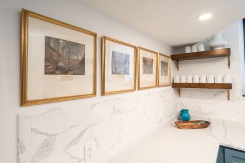 A corner of a kitchen featuring framed artwork, white marble backsplash, wooden shelves, and decorative items.