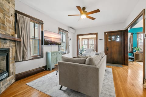 Cozy living room featuring a gray sofa, stone fireplace, and a view into a sunny room.