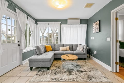 A living room featuring a gray sectional sofa, large windows with white curtains, and a wooden coffee table.