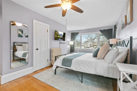 A beautifully decorated bedroom featuring a bed with white linens, a mirror, and large windows with green curtains.