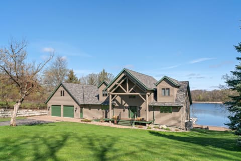 Lakeside house surrounded by green lawn and trees, with a scenic view of a lake and blue sky.