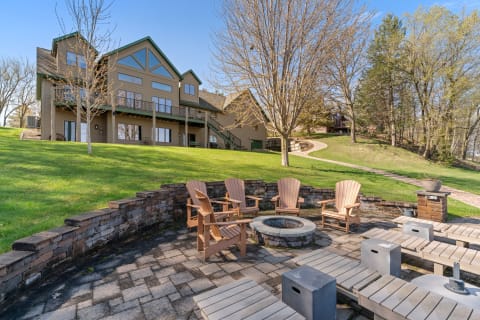 Outdoor patio area with wooden chairs around a fire pit, set against a grassy hill and a large house.