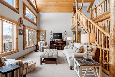 A spacious lodge-style living room featuring a high ceiling, large windows, and rustic furnishings.