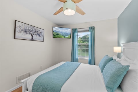 Cozy bedroom with blue accents, a framed tree artwork, and a mounted TV displaying a landscape.
