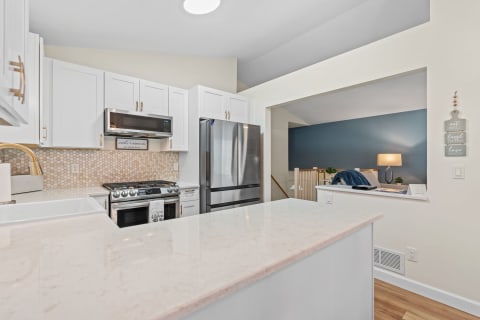 Modern kitchen features white cabinets, light stone countertop, and stylish appliances leading to a cozy living area.