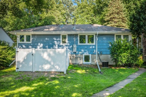 A single-story blue house with a white gate, green grass, and trees in the background.