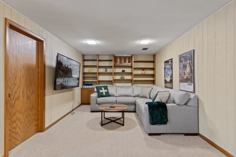A cozy living room in a basement featuring a gray sectional sofa, wooden shelves with decorative items, and movie posters on the walls.