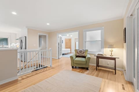 An interior view showing a green armchair, a wooden table with a lamp, and an open doorway to a kitchen.