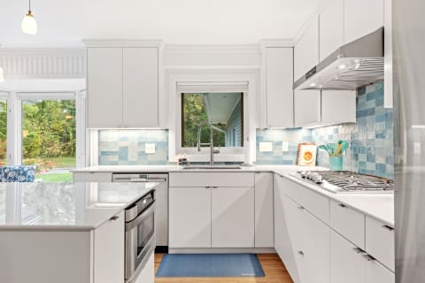 A bright modern kitchen featuring white cabinets and a blue tile backsplash with natural light from large windows.