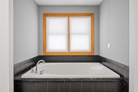 A spacious bathroom with a white bathtub and gray walls, illuminated by natural light from two windows.